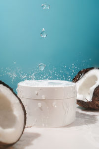 Two halves of coconut and a jar of cosmetics on a blue background with water drops.