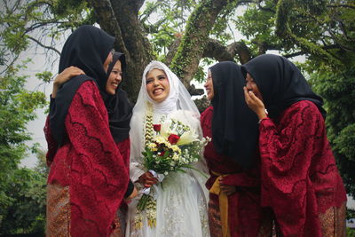 Cheerful bride and bridesmaids standing against tree