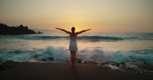 Rear view of woman with arms outstretched standing at beach against sky during sunset