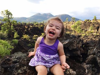 Portrait of cheerful girl sitting on rock against sky