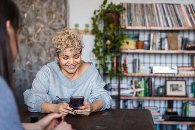 Smiling woman text messaging on smart phone while sitting with friend at home