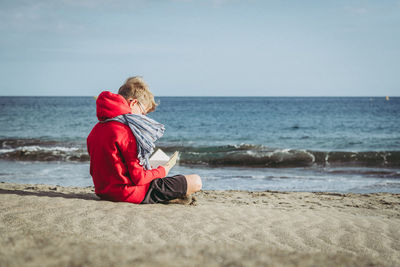 Side view of boy reading book while sitting at beach against sky during sunny day