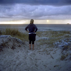 Full length rear view of woman with hands on hip looking at sea against cloudy sky during sunset