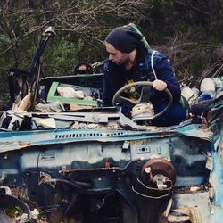 Man sitting in abandoned damaged car at garbage dump