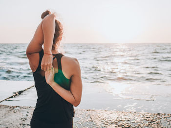 Woman doing yoga at beach against sky during sunset