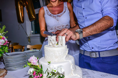 Midsection of couple cutting wedding cake on table