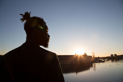 Thoughtful man standing by lake and city against clear sky during sunset