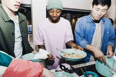 Young man with noodles while roommate cleaning dishes at sink in college dorm