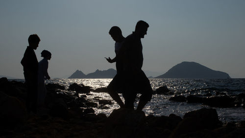 Silhouette people on rock by sea against sky