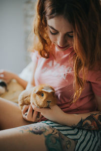 Young woman in pajamas playing with guinea pig and relaxing on bed in bedroom