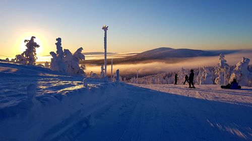 People skiing on snow covered field against sky during sunset