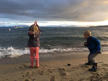 Siblings playing on seashore against cloudy sky during sunset