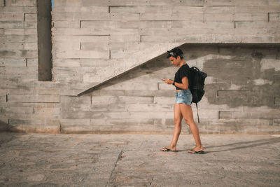 Full length side view of woman using mobile phone walking against wall outdoors