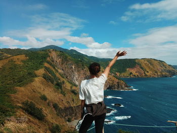 Rear view of woman waving hand while standing on cliff by sea against sky