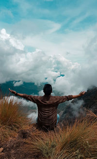 Rear view of man with arms outstretched sitting on mountain against cloudy sky