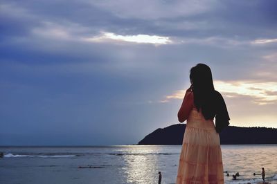 Rear view of woman standing at beach against cloudy sky during sunset