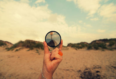 Cropped hand holding mirror at beach against cloudy sky