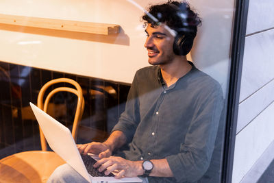 Young smiling hispanic man in black shirt listening to music in wireless headphones and browsing data on netbook while sitting behind cafe window and working on freelance project