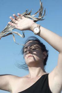Low angle view of woman holding leaf while shielding eyes against blue sky during sunny day