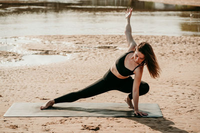 Tranquil female practicing yoga with closed eyes in side lunge with arms extended pose while standing on mat on beach in summer