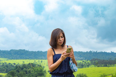 Woman using phone while standing on green landscape against cloudy sky