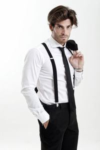 Portrait of man wearing suspenders standing with hands in pockets against white background