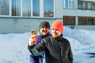 Man in warm clothing taking selfie with friend through mobile phone