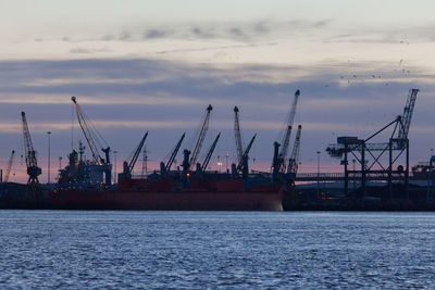 Cranes at commercial dock against cloudy sky at dusk