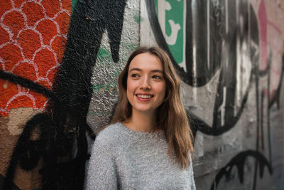 Positive young female leaning on wall with colorful creative graffiti while standing in urban area and looking away