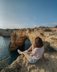 Side view of woman sitting on rock against sky