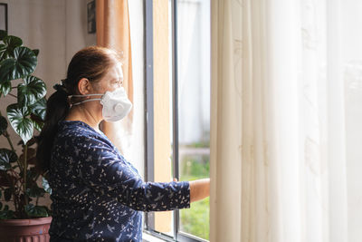 Woman wearing mask looking through window at home