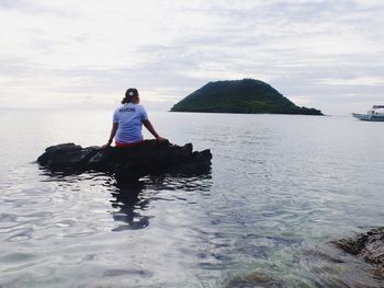 Rear view of woman sitting on rock in sea against cloudy sky