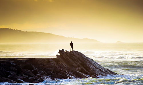 Silhouette man standing on rock by sea against sky during sunset
