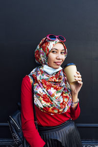 Smiling woman holding disposable coffee cup against black wall during covid-19