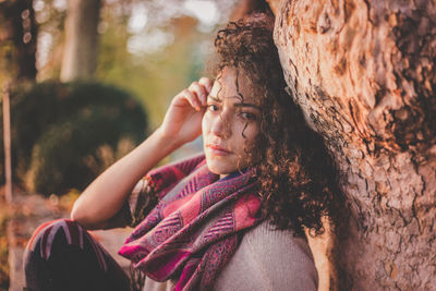 Portrait of young woman sitting by tree trunk at park