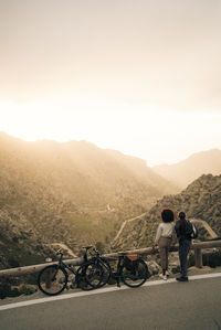 Friends looking at mountains while standing roadside with bicycles at sunset on vacation