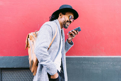 Side view of glad hispanic man in trendy clothes with backpack holding hand in pocket and smiling while leaning on pink and gray building wall and sending audio message via smartphone