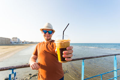 Portrait of mid adult man holding sunglasses at beach against sky