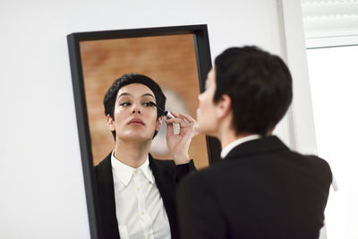 Woman applying mascara while standing against mirror