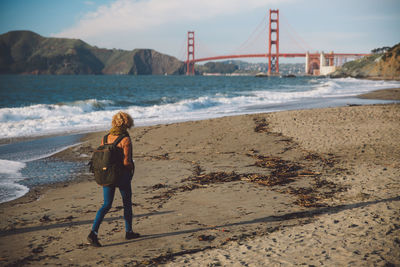 Rear view of mid adult woman with backpack standing at beach against sky