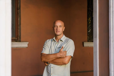 A balding middle-aged man in a plaid shirt stands against a painted wall