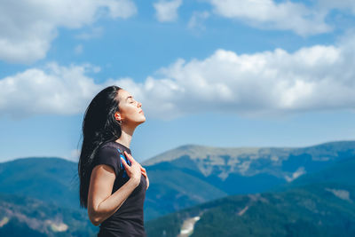 Side view of mid adult woman with eyes closed standing on mountain against cloudy sky