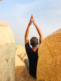 Rear view of man with arms raised meditating while sitting on rock against sky