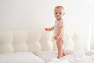 Cute baby girl in bodysuit stand holding bed frame, learning to walk on white bedding on bed