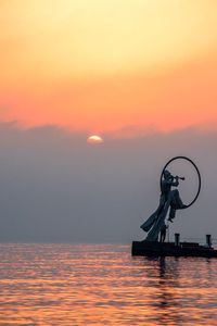 Silhouette sculpture by sea against orange sky