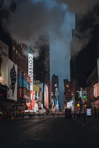 Illuminated city street by buildings against sky at night