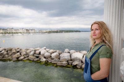 Mature woman looking away while standing by lake against cloudy sky