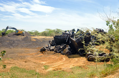 Earth mover on field besides wreckage of abandoned train against sky