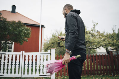 Man holding bouquet of flowers while using mobile phone against house