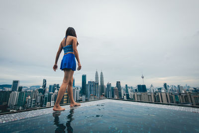 Rear view of woman standing by swimming pool against buildings in city